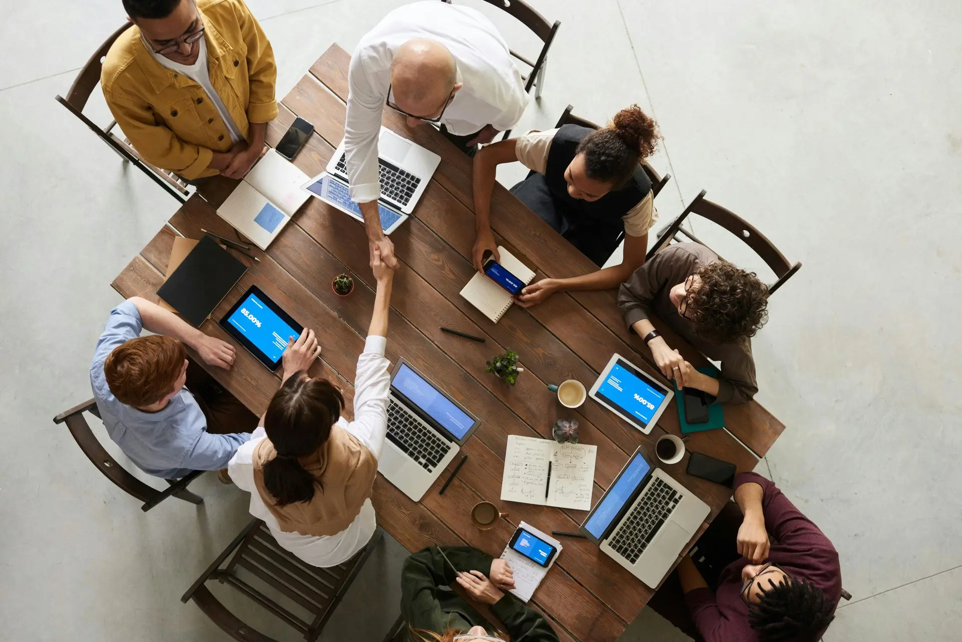 8 people in a meeting with the tablets and laptops out. Two people are shaking hands from across the table.