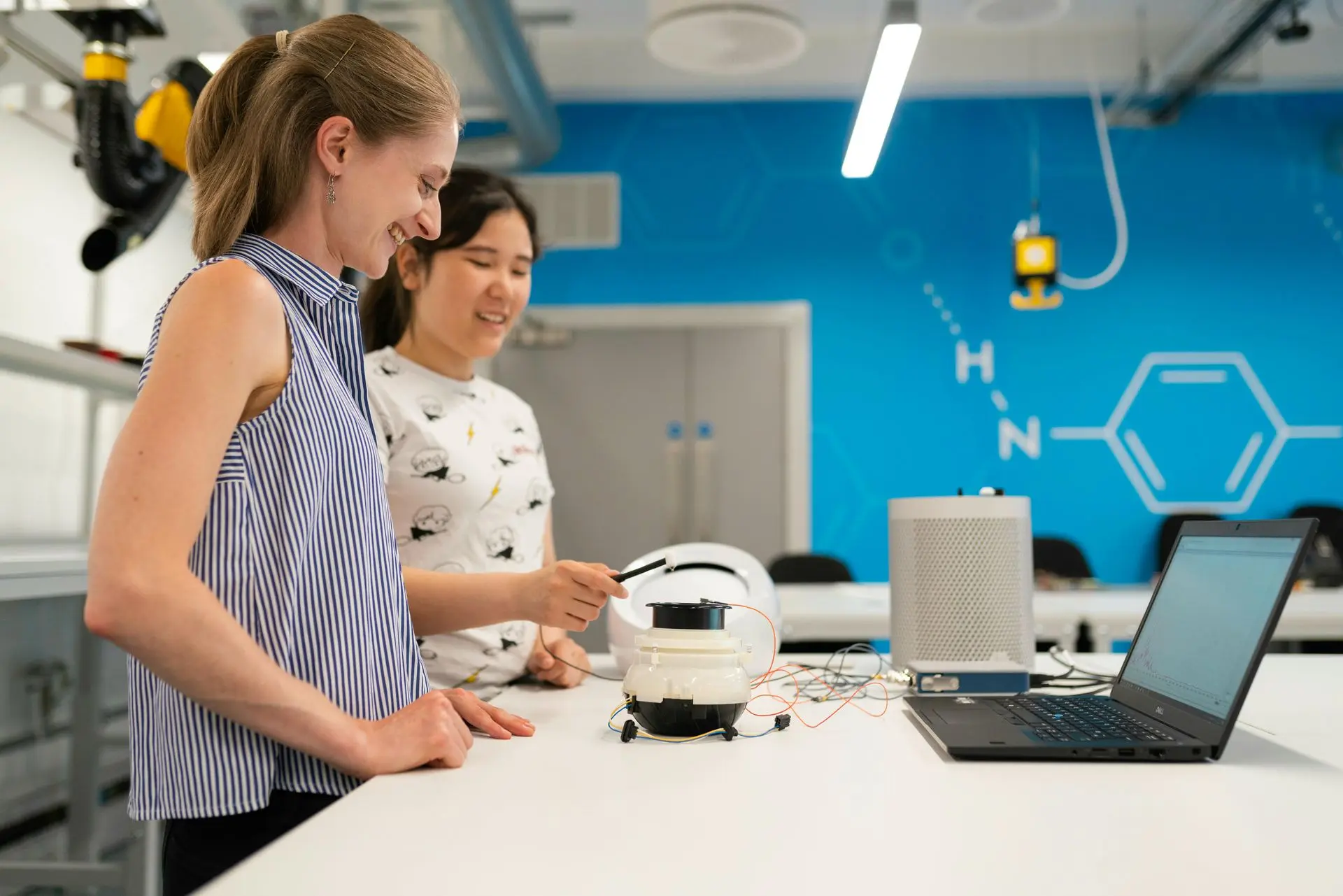 Two women in a lab setup with a laptop and a representation of a chemical formula on the back wall